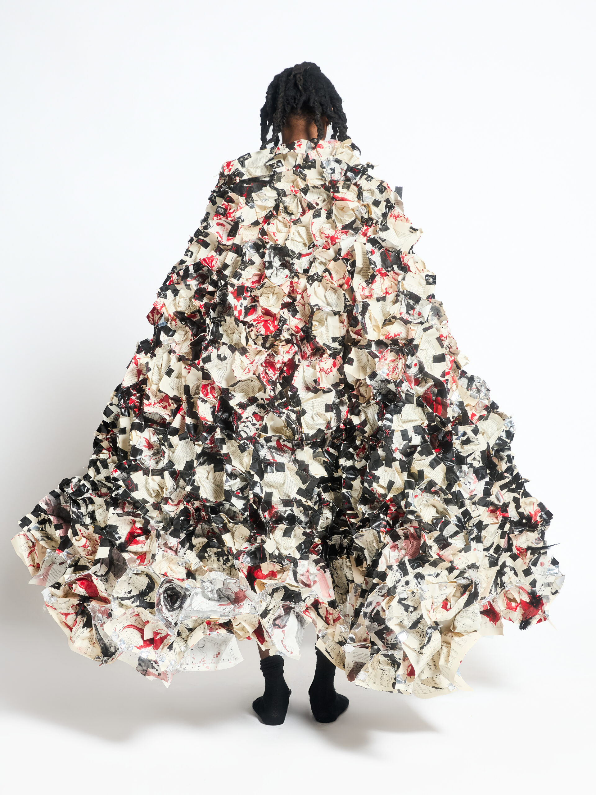 A person stands in a voluminous, abstract gown made of crumpled paper with black, white, and red patterns, against a plain white background.
