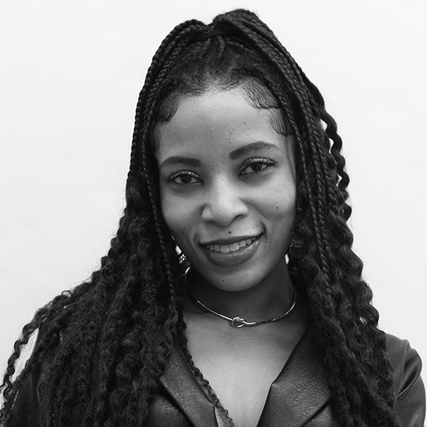 A person with long, braided hair smiles softly while wearing a choker and a textured outfit, set against a plain white background.