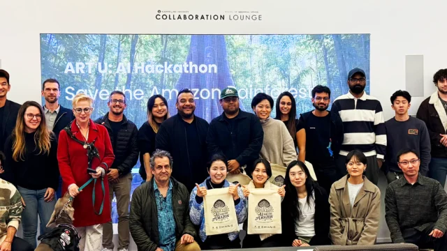 A group of diverse individuals stands in a room labeled "COLLABORATION LOUNGE." They hold bags with "AI FOR THE AMAZON" printed. The screen behind reads, "ART U: AI Hackathon Save the Amazon rainforest."