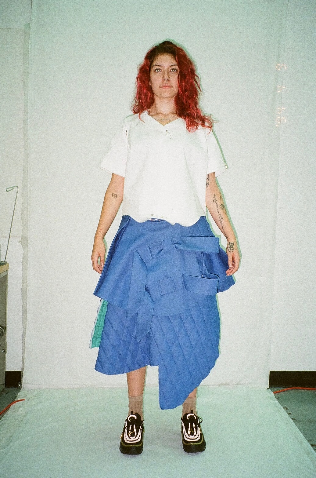 A person stands wearing a white shirt and a blue, quilted skirt with layered fabric. The background is a plain white backdrop, suggesting a studio setting.