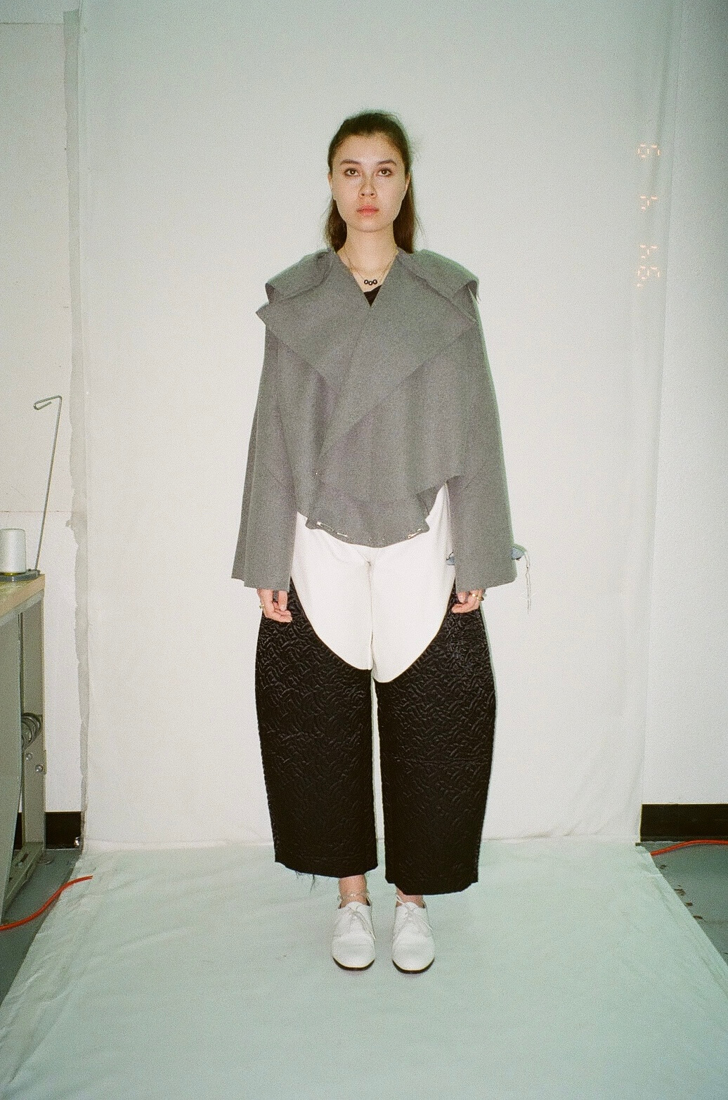 A person stands in a studio setting, wearing a gray layered jacket, white and black wide-legged pants, and white shoes against a white backdrop. Text is not present.