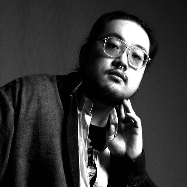 Person wearing glasses and a nose ring, resting their chin on their hand, looking directly at the camera. The setting is a grayscale, studio background.