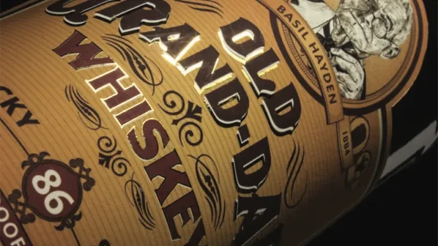 A whiskey bottle label displays "Old Grand-Dad Whiskey" with "86 Proof" beneath. The background features ornate designs and a portrait labeled "Basil Hayden." The label is angled against a dark background.